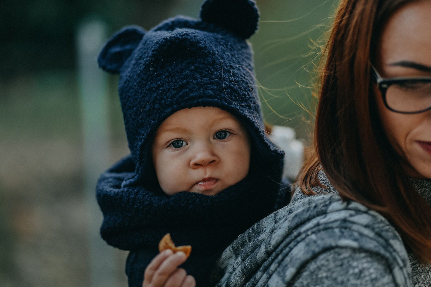 Dziecko siedzi w chuście na plecach u mamy. Ma czapkę z uszami misia