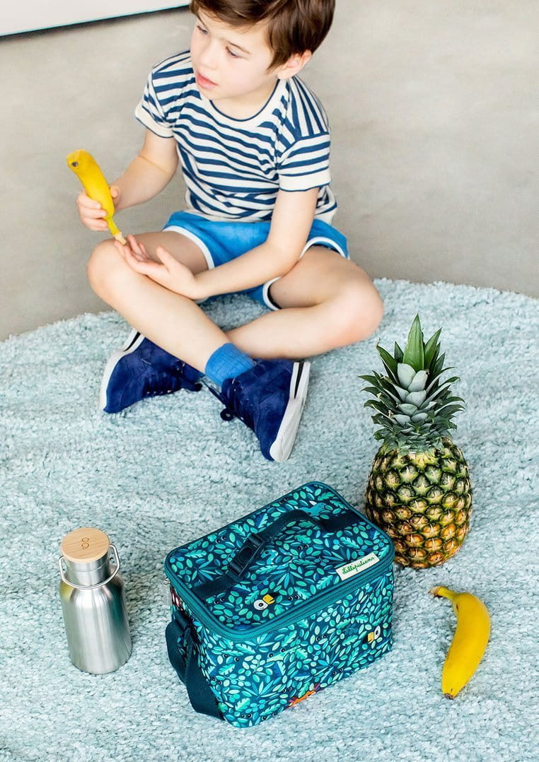 Chłopiec siedzi po turecku na podłodze, przed nim jest termos, torba termiczna, ananas i banan