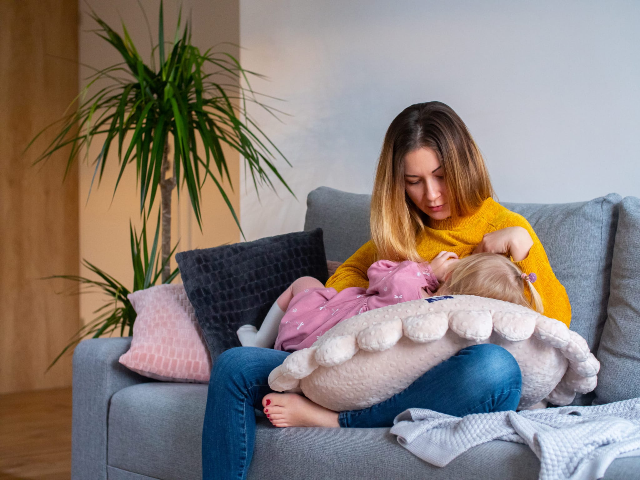 Mama siedzi na kanapie, na nogach ma poduszkę do karmienia i oparte o nią dziecko