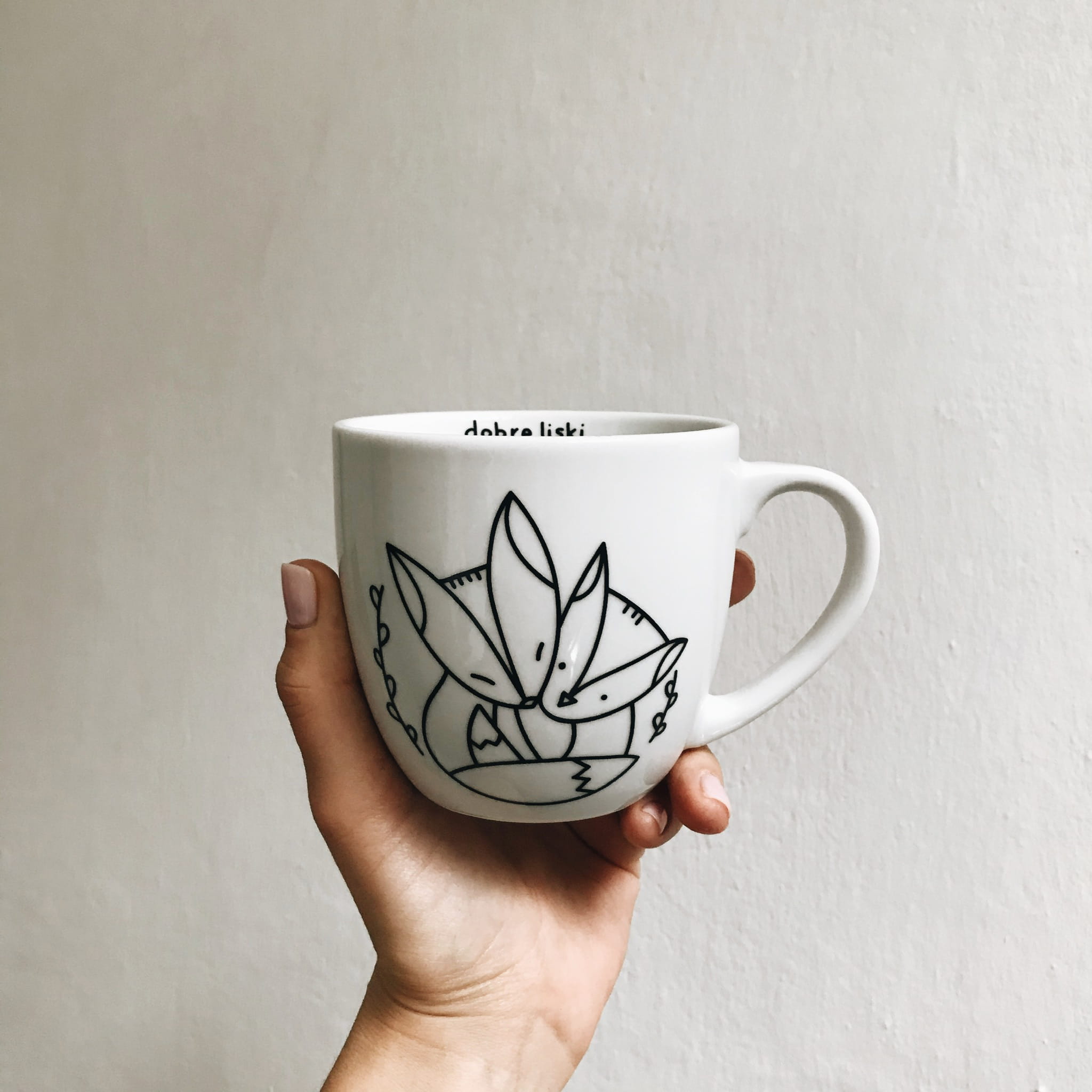 Biały kubek trzymany w dłoni. Na białej porcelanie naniesiony jest czarny, geometryczny kontur lisków