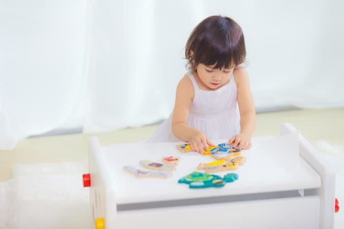 Dziewczynka z ciemnymi włosami w białym ubranku pochyla się nad stołem i układa puzzle